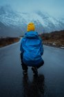 Vista posterior de una persona relajada irreconocible en ropa de abrigo azul y sombrero de gorro amarillo brillante agachado en el camino de asfalto que va a las montañas nevadas de niebla en Lofoten - foto de stock
