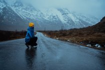 Side view of relaxed woman in blue warm clothes and bright yellow beanie hat enjoying life while crouched looking away on asphalt road going to snowy foggy mountains in Lofoten — Stock Photo