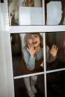 Мила дівчинка в повсякденному одязі дивиться на камеру і посміхається, стоячи за вікном вдома і торкаючись скла — стокове фото