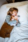 Excited little girl hugging plush toy and laughing while lying on soft bed near fairy lights at home — Stock Photo