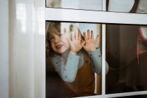 Petite fille mignonne en vêtements décontractés regardant la caméra et souriant tout en se tenant derrière la fenêtre à la maison et touchant le verre — Photo de stock