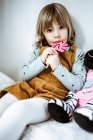 Nettes kleines Mädchen in lässiger Kleidung schaut in die Kamera und saugt Lutscher, während es auf einem bequemen Bett in der Nähe von Spielzeug zu Hause sitzt — Stockfoto