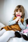 Menina bonito em roupas casuais olhando para a câmera e chupando pirulito enquanto sentado na cama confortável perto de brinquedo em casa — Fotografia de Stock