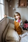 Menina adorável em roupas casuais chupando pirulito doce e olhando para fora da janela enquanto descansa no quarto acolhedor em casa — Fotografia de Stock