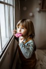 Adorable niña en ropa casual chupando piruleta dulce y mirando por la ventana mientras descansa en la acogedora habitación en casa - foto de stock