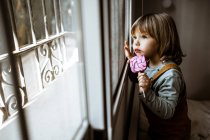 Чарівна дівчинка в повсякденному одязі смокче солодкий льодяник і дивиться у вікно, відпочиваючи в затишній кімнаті вдома — стокове фото