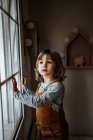 Adorabile bambina in abiti casual guardando fuori dalla finestra mentre riposava in camera accogliente a casa — Foto stock