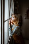 Liebenswertes kleines Mädchen in lässiger Kleidung, das aus dem Fenster schaut, während es sich zu Hause in einem gemütlichen Zimmer ausruht — Stockfoto