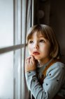 Adorable petite fille en vêtements décontractés regardant par la fenêtre tout en se reposant dans une chambre confortable à la maison — Photo de stock