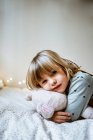 Eccitato bambina abbracciando peluche giocattolo e ridendo mentre sdraiato sul letto morbido vicino alle luci delle fate a casa — Foto stock