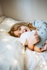 Eccitato bambina abbracciando peluche giocattolo e ridendo mentre sdraiato sul letto morbido vicino alle luci delle fate a casa — Foto stock