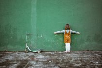 Back view of anonymous little girl with outstretched arms standing near weathered green wall and kick scooter and counting while playing hide and seek — Stock Photo