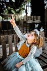 Красивая маленькая девочка в сказочном костюме сидит на потрепанной деревянной скамейке и смотрит в камеру, проводя время во дворе — стоковое фото