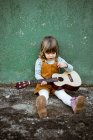 Little girl with ukulele sitting on rough ground near kick scooter against weathered green wall on street — Stock Photo
