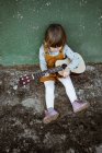 Kleines Mädchen mit Ukulele sitzt auf unwegsamem Boden neben Tretroller gegen verwitterte grüne Mauer auf der Straße — Stockfoto