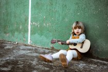 Little girl with ukulele sitting on rough ground near kick scooter against weathered green wall on street — Stock Photo