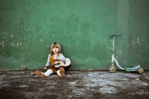Kleines Mädchen mit Ukulele sitzt auf unwegsamem Boden neben Tretroller gegen verwitterte grüne Mauer auf der Straße — Stockfoto