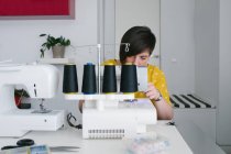Focalisé brunette femme adulte souriant et en utilisant une machine à coudre pour faire du vêtement tout en travaillant à la maison atelier — Photo de stock