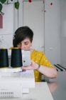 Konzentrierte brünette erwachsene Frau lächelt und verwendet Nähmaschine, um Kleidungsstücke zu machen, während sie in der heimischen Werkstatt arbeitet — Stockfoto