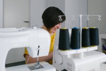 Mulher adulta morena feliz sorrindo e usando máquina de costura para fazer roupas enquanto trabalhava em oficina em casa — Fotografia de Stock