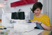 Щаслива брюнетка доросла жінка посміхається і використовує швейну машину для виготовлення джинсового одягу під час роботи в домашній майстерні — стокове фото