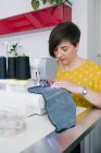 Орієнтована брюнетка доросла жінка посміхається і використовує швейну машину для виготовлення джинсового одягу під час роботи в домашній майстерні — стокове фото