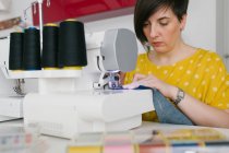 Сфокусированная брюнетка взрослая женщина улыбается и использует швейную машинку, чтобы сделать джинсовую одежду во время работы в домашней мастерской — стоковое фото