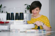 Mulher adulta morena feliz sorrindo e usando máquina de costura para fazer roupas jeans enquanto trabalhava em oficina em casa — Fotografia de Stock