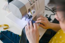 From above cropped unrecognizable brunette adult woman using sewing machine to make denim garment while working in home workshop — Stock Photo