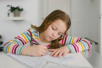Menina focada em t-shirt listrada escrevendo em notebook enquanto sentado à mesa e fazendo lição de casa em casa — Fotografia de Stock