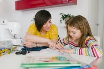 Happy adult woman smiling and helping girl with homework assignment while sewing garment at home — Stock Photo