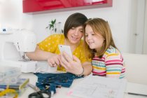 Femme adulte joyeuse souriant et prenant selfie avec la fille tout en étant assis à la table et travaillant dans l'atelier de couture à la maison — Photo de stock