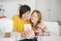 Fröhliche erwachsene Frau lächelt und macht Selfie mit Mädchen, während sie am Tisch sitzt und zu Hause in der Schneiderei arbeitet — Stockfoto