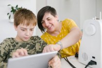 Mulher adulta ajudando menino navegando tablet moderno juntos enquanto sentados à mesa em casa — Fotografia de Stock