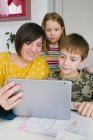 Erwachsene Frau und Kinder surfen gemeinsam am modernen Tablet, während sie zu Hause am Tisch sitzen — Stockfoto