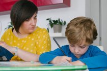 Сосредоточенный мальчик делает домашнее задание, сидя рядом со взрослой женщиной, шьет одежду дома — стоковое фото