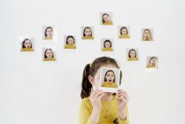 Cute little girl in yellow dress hiding face behind own picture while standing against wall with photos demonstrating various emotions — Stock Photo