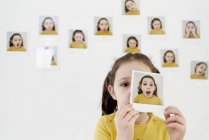 Симпатична маленька дівчинка в жовтій сукні ховається обличчям за власною картиною, стоячи на стіні з фотографіями, демонструючи різні емоції — стокове фото