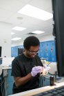 Afro-Américain dans des gants en latex en utilisant un miroir de bouche et une sonde pour vérifier les fausses dents tout en travaillant dans un laboratoire moderne — Photo de stock