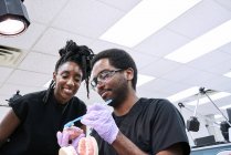 Angle bas de femme afro-américaine heureuse avec dreadlocks et homme barbu souriant et brossant de fausses dents pendant le travail en laboratoire — Photo de stock