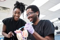 Bajo ángulo de mujer afroamericana feliz con rastas y hombre barbudo sonriendo y cepillando dientes postizos durante el trabajo en el laboratorio - foto de stock