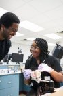 Délicieuse femme afro-américaine avec des tresses souriantes et utilisant un miroir de bouche et une sonde pour montrer à ses collègues de fausses dents pendant le travail en laboratoire — Photo de stock
