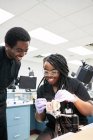 Hembra afroamericana encantada con trenzas sonriendo y usando espejo bucal y sonda para mostrar dientes falsos de compañeros de trabajo durante el trabajo en el laboratorio - foto de stock