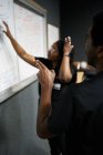 Чорний чоловік і жінка з косами читають і обговорюють нотатки на дошці під час роботи в сучасній лабораторії разом — стокове фото