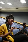 Знизу чорна жінка з дредлоками посміхається і використовує планшет, сидячи в освітленому лекційному залі під час уроку — стокове фото