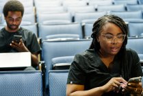 Black woman with braids and African American man sitting in auditorium and browsing smartphones during lesson in auditorium — Stock Photo