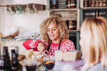 Mulher adulta feliz com cabelo encaracolado conversando com o cliente enquanto vende queijo na loja local de alimentos delicatessen — Fotografia de Stock
