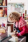 Mujer adulta en delantal rebanando queso fresco mientras trabaja en una acogedora tienda de comida local - foto de stock