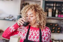 Glücklich lockiges Haar blonde Verkäuferin Barkeeper trinken ein Glas Tasse mit Wein während der Arbeit in der lokalen Feinheit Lebensmittelgeschäft — Stockfoto
