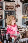 Friendly adult woman with curly hair leaning on shelf and looking away while working in cozy local delicatessen food shop — Stock Photo